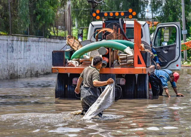 Workers wading through floodwaters near a truck, managing a city crisis after heavy rain. How to start water restoration business without skipping licensing